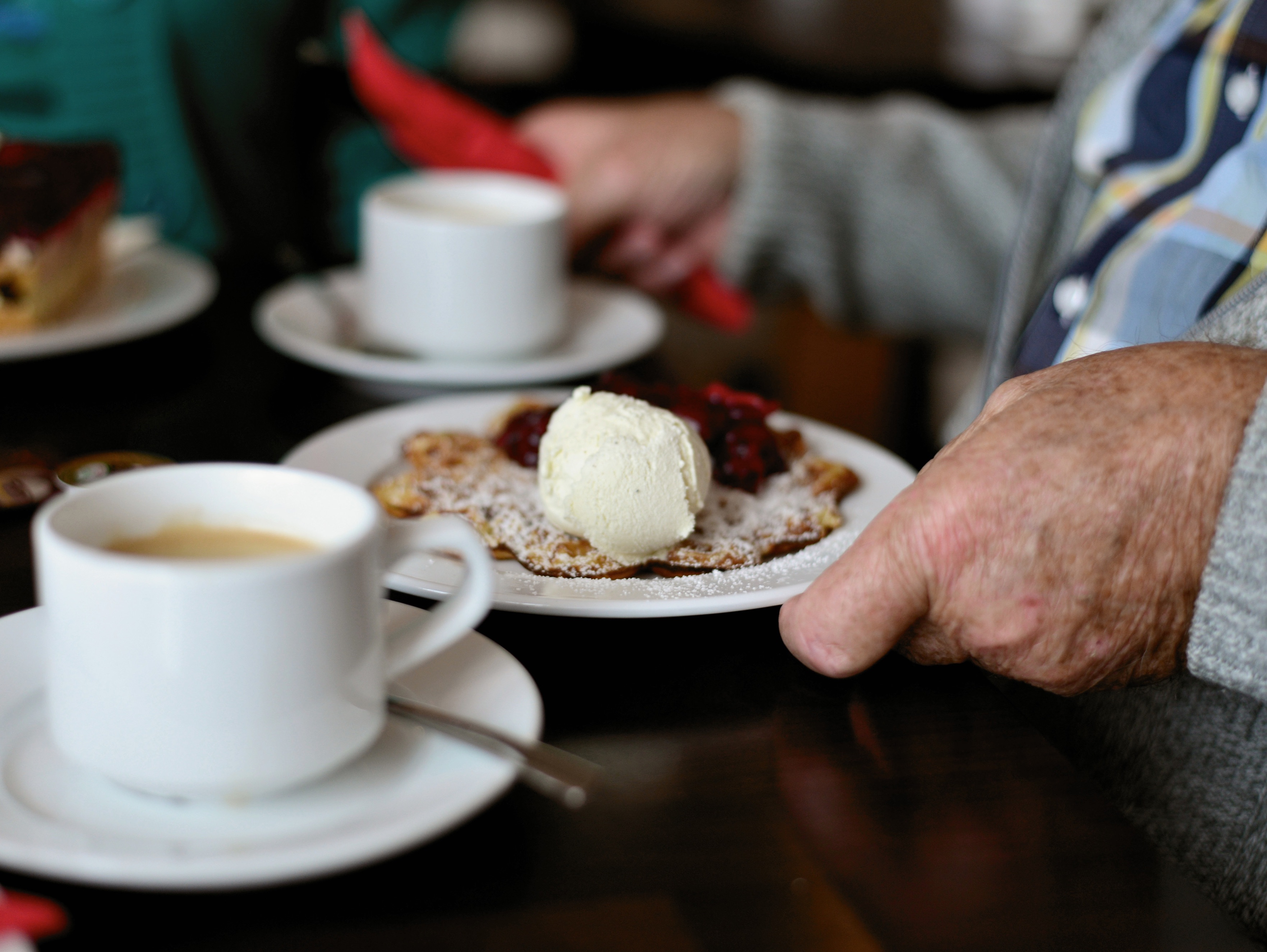 Die Hand eines älteren Mannes, der einen Teller mit einer Waffel hält, vor ihm eine Kaffeetasse auf dem Tisch
