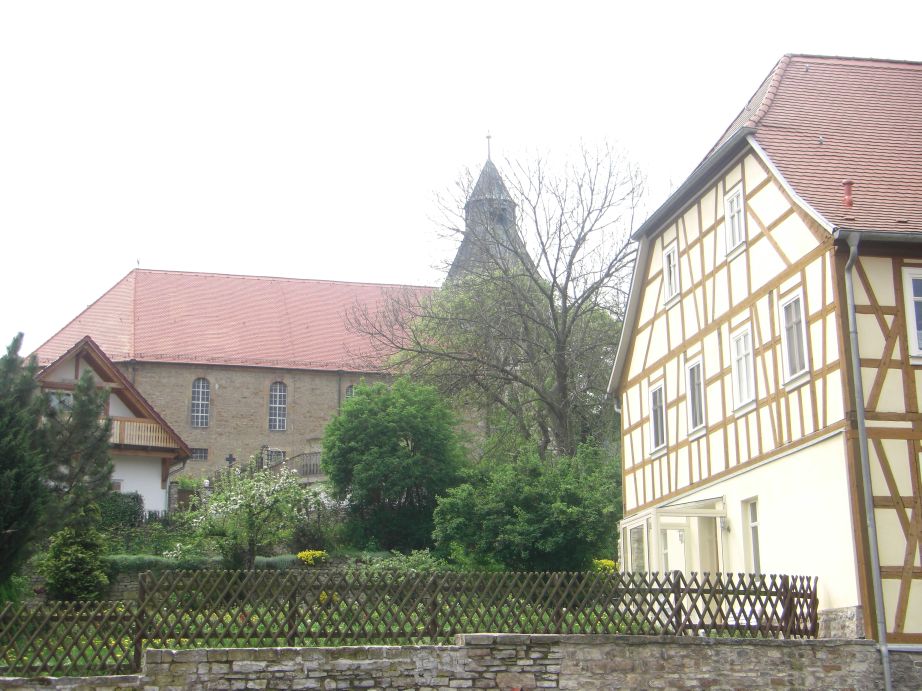 grün bewachsener Garten hinter einem Zaun, daneben ein Fachwerkhaus, im Hintergrund eine Kirche