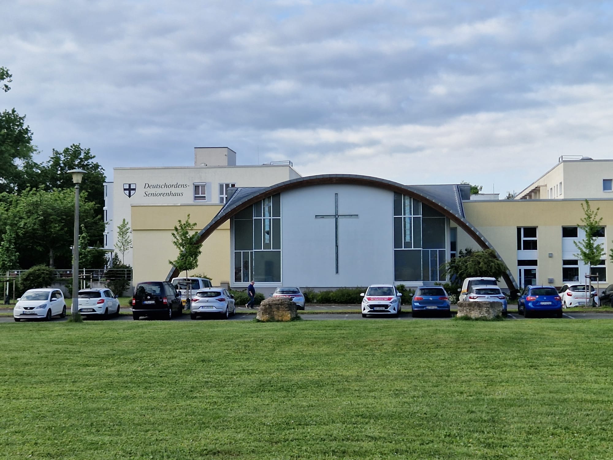 Gebäudekomplex aus Beton mit großem Kreuz an der Vorderseite, davor parkende Autos und grüne Wiese