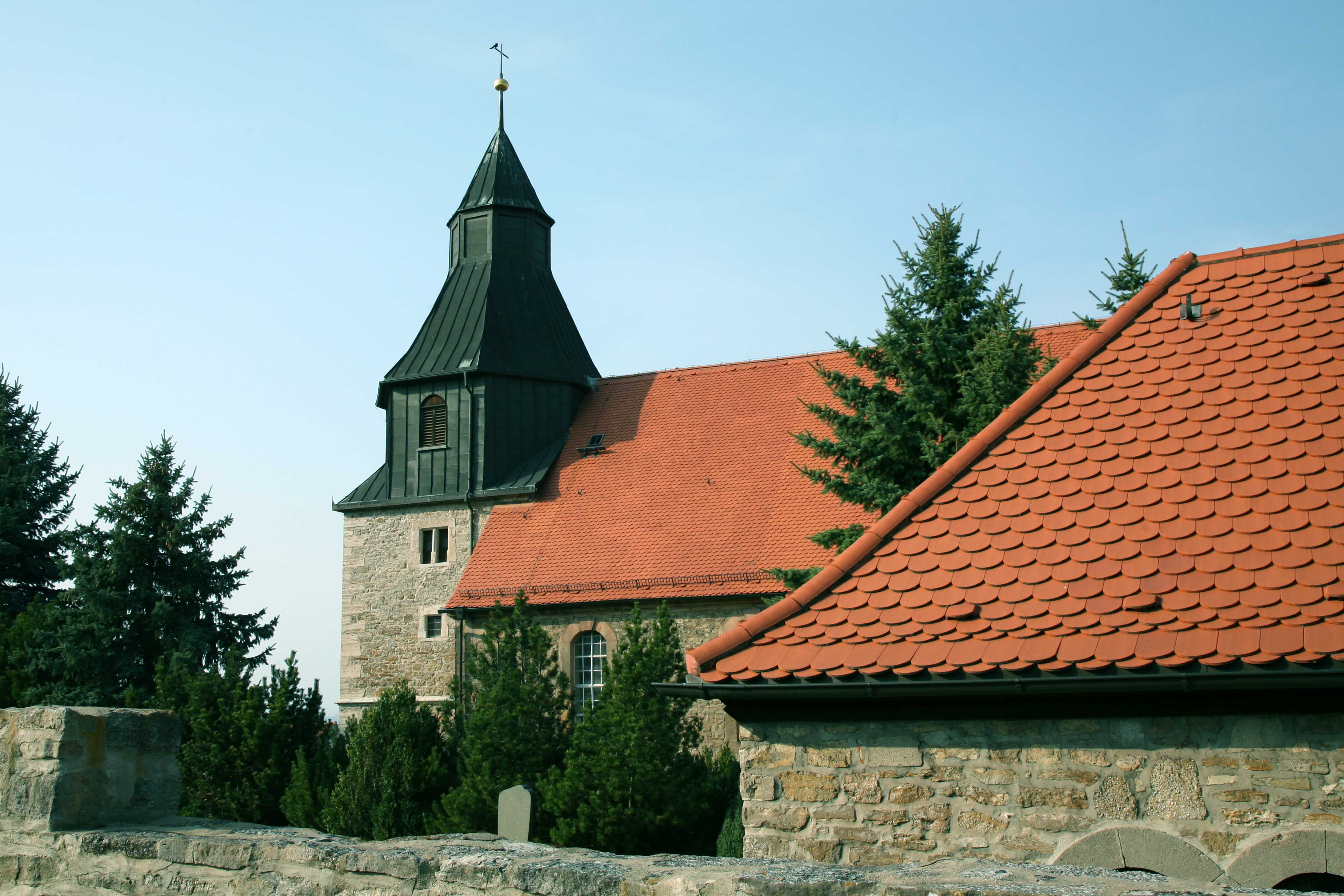 Kirche aus grauen Steinen mit roten Dachziegeln und einem Turm mit schwarzem Metalldach