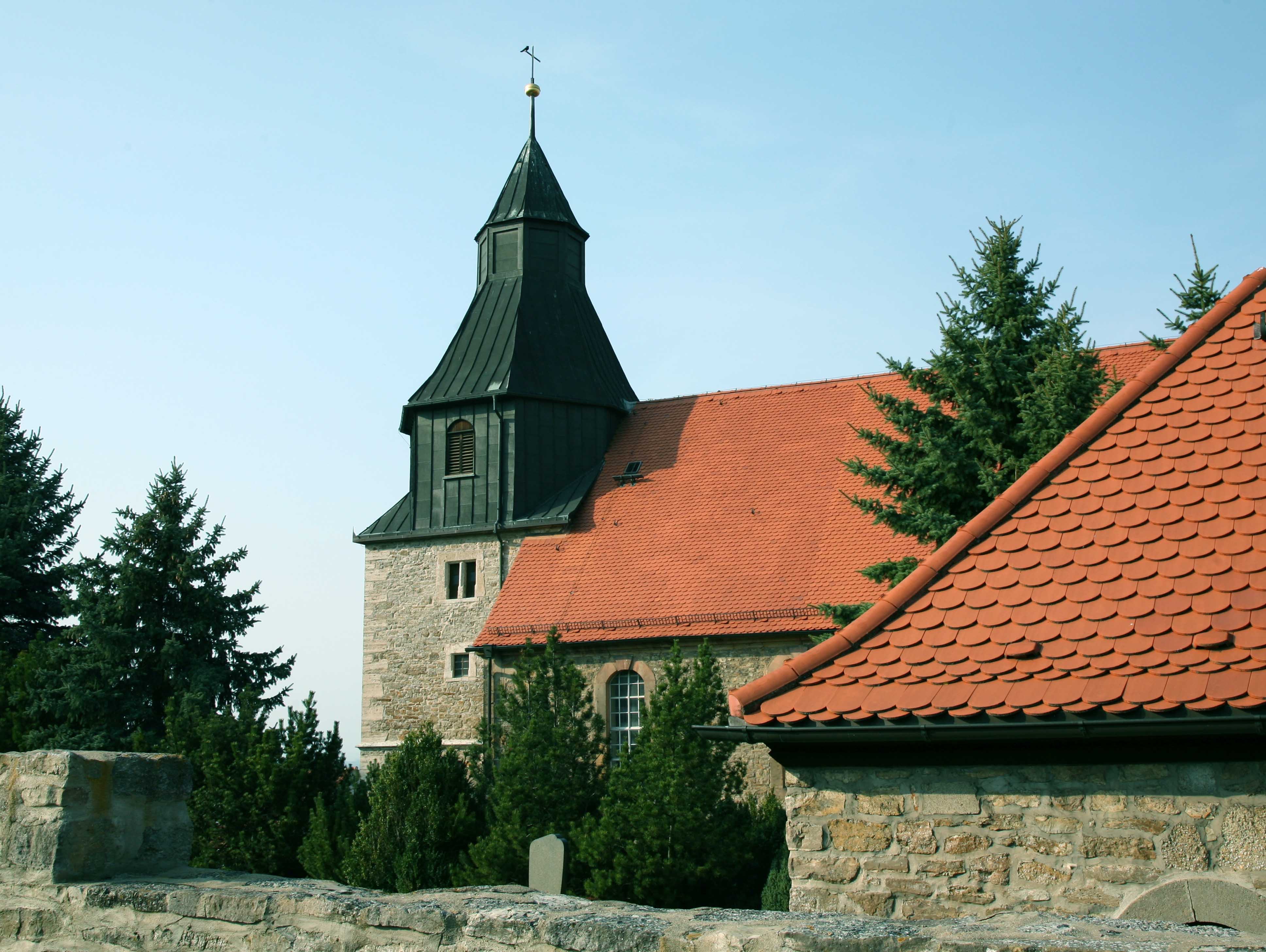 Kirche aus grauen Steinen mit roten Dachziegeln, auf dem Kirchturm ein schwarzes Metalldach